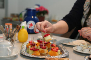 Foto: Ein Tisch mit Obstspießen und anderen Lebensmitteln sowie ein Teller mit den Händen einer Frau, die isst.